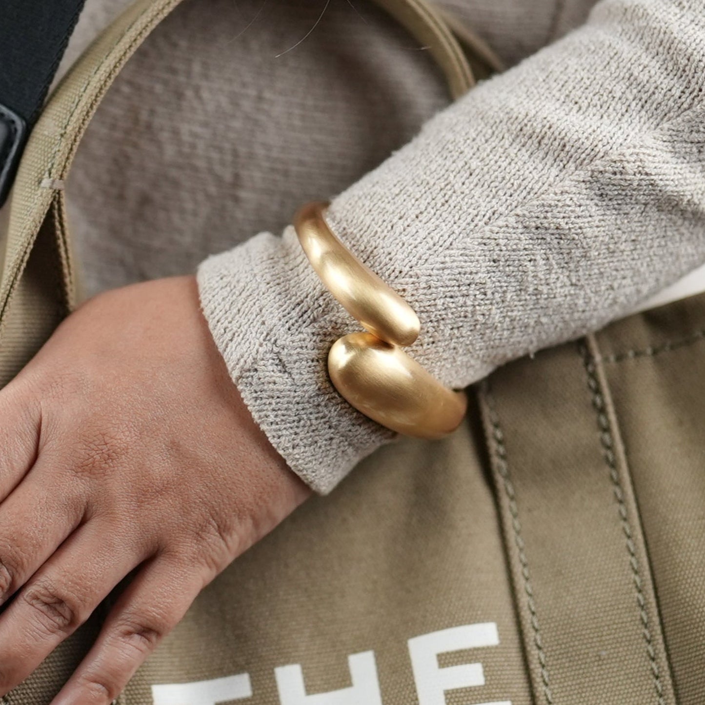 Elegant matt titanium finish gold-toned statement bangle bracelet by Adiva Jewelry, styled on a woman's wrist with a neutral-toned sweater and canvas tote bag — a bold yet minimal accessory perfect for everyday sophistication