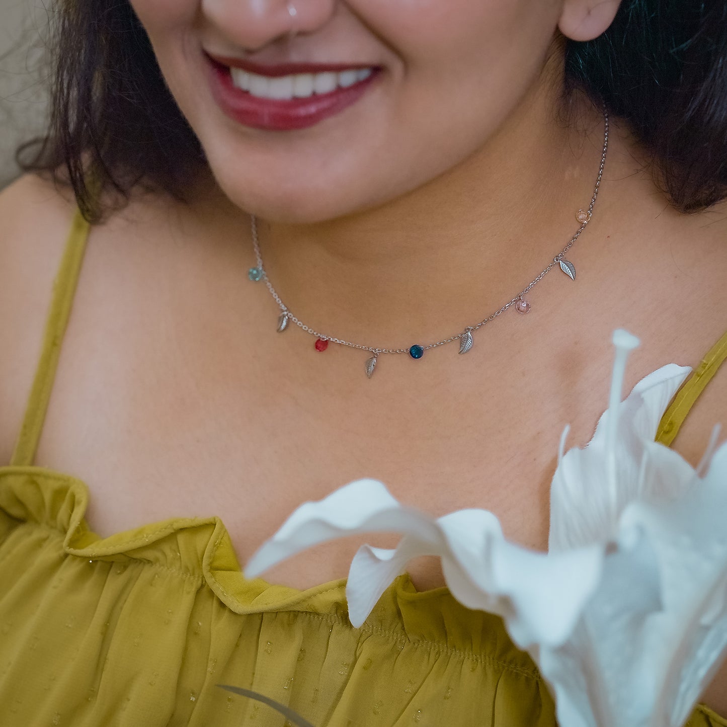 Close-up of a woman wearing a delicate necklace made of leaf and small colored stone charms touching her chest. Necklace is made of sterling silver and is by Adiva