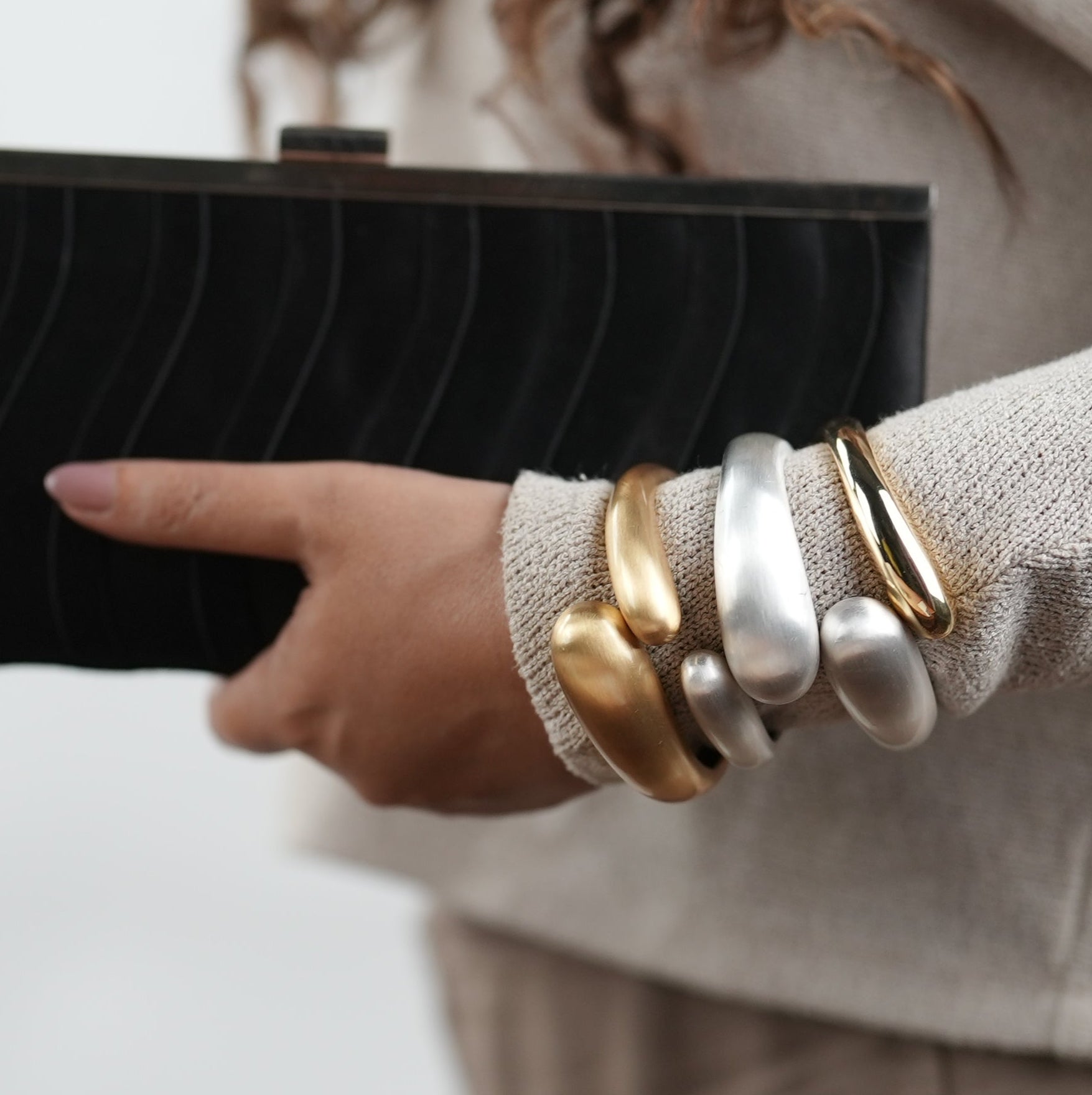 Stacked statement cuff bracelets in gold and matte silver finish, worn on wrist with beige knit top and black clutch – modern chunky metal bangles by Adiva Jewelry for bold fashion styling